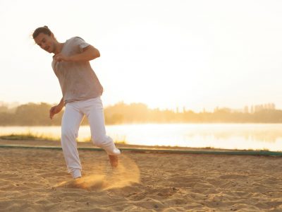 Concept about people, lifestyle and sport. Capoeira on the beach, near lake in the park one performer, at sunrise. Casual athletic man in white pants making capoeira movements on the beach